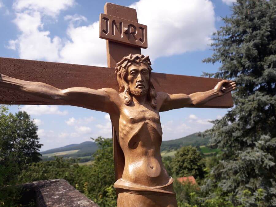 Vor einem weiss-blauen Himmel steht ein Holzkreuz auf dem Jesus aufrecht als Sieger nach vorn blickt.
